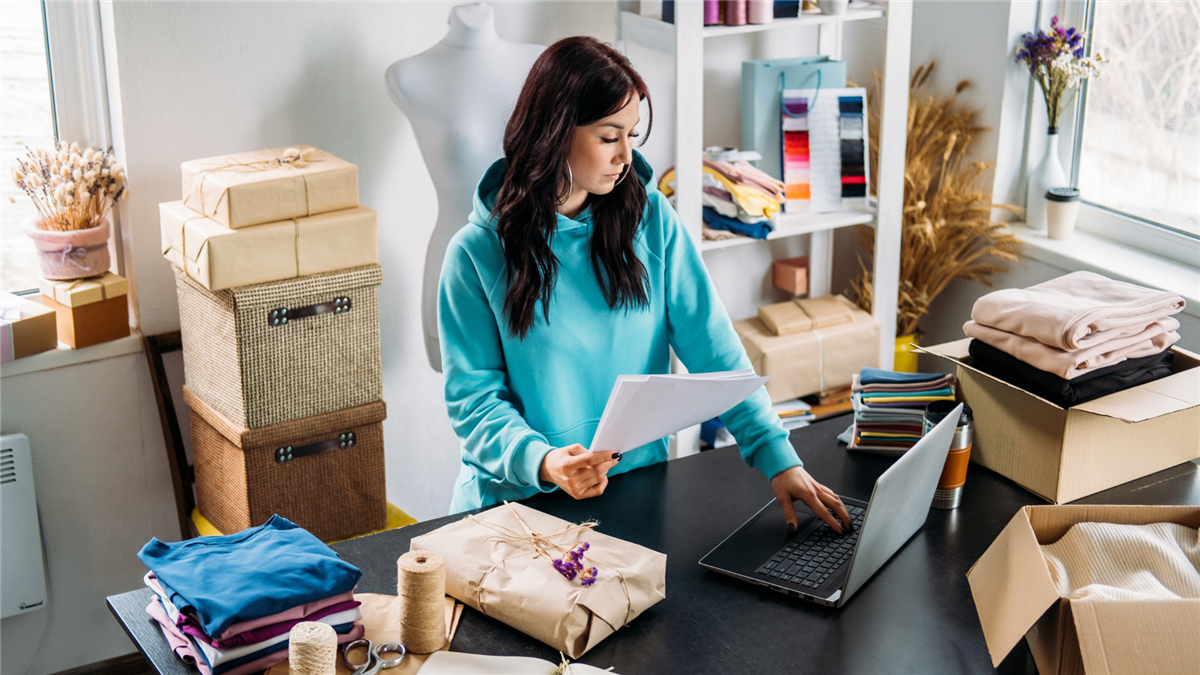 Woman in blue jumper stands at computer processing orders, surrounded by packages.